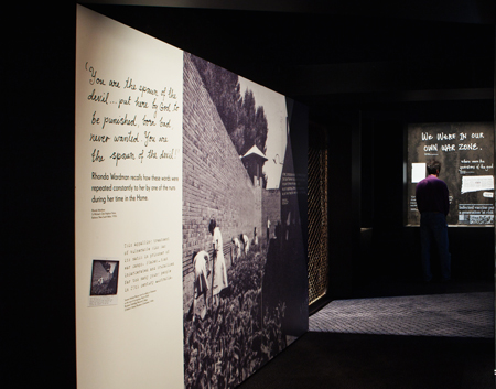 Large exhibition wall panel at left, showing girls working in the garden, watched over by officers, 1965